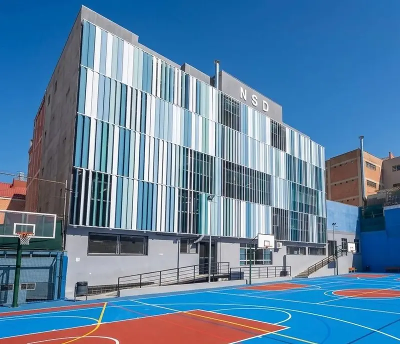 Patio y pista del colegio, con la canasta y la fachada de lamas azules y verdes al fondo
